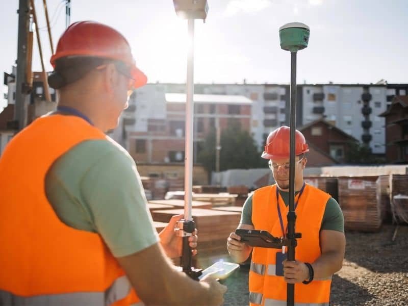 Survey crew establishing construction control points beside a commercial corridor with retail buildings, parking areas, and road access.